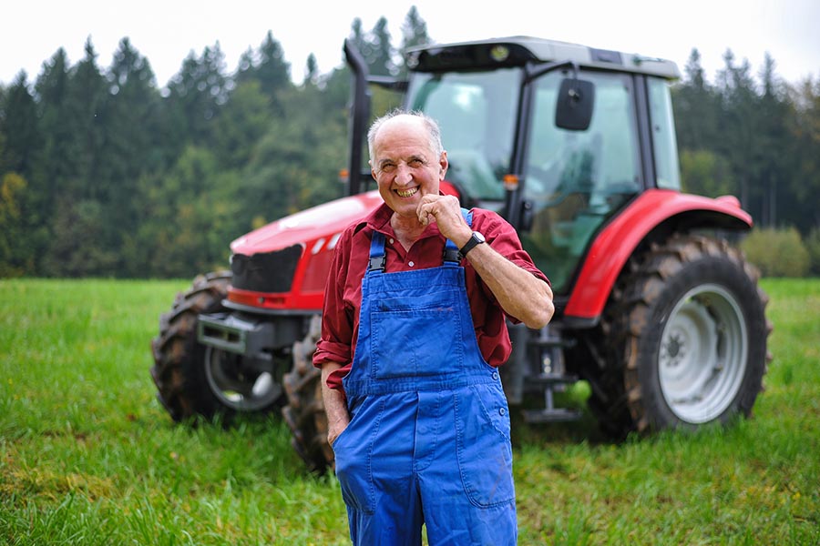 Business Insurance - Farmer Smiles and Poses in Front of His Tractor in a Green Field, Wearing Red Shirt and Blue Overalls