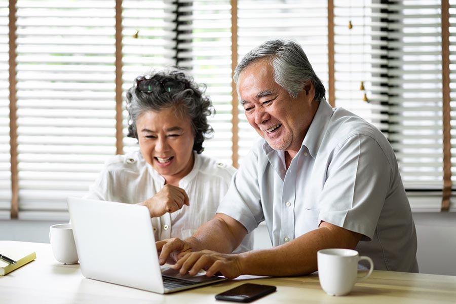 Client Center - Senior Couple Laughs as They Use a Laptop at Their Kitchen Table, Coffee and Cell Phone Nearby